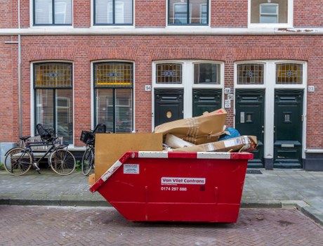 A large red skip bin filled with assorted waste materials parked on a sidewalk in front of a row of brick terraced houses in Shoreditch. The waste in the bin includes cardboard boxes, paper, and plastic packaging. Two bicycles are leaning against the house wall on the left side of the image, and the houses feature tall, narrow windows with yellow and white frames, along with dark green doors. The scene appears to be in an urban residential area, with the skip used for a local clearance, possibly managed by Office Clearance Shoreditch, given the context of rubbish removal services. The pavement is made of concrete, and the background shows a clear, bright day with natural light illuminating the scene.