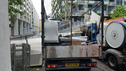 A flatbed truck parked on a city street in Shoreditch, loaded with various discarded office furniture and debris. Visible items include white and beige office chairs, broken or damaged desks, and assorted packaging materials. The truck's metal surface shows signs of rust and wear, with some parts covered by a wire mesh barrier. In the background, modern apartment buildings and street infrastructure such as traffic lights and pedestrian crossings can be seen. The scene is captured in daylight with typical urban lighting, emphasizing the contrast between the clean lines of the surrounding buildings and the utilitarian, cluttered appearance of the rubbish on the truck, which Office Clearance Shoreditch likely services for local office clearance and rubbish removal.