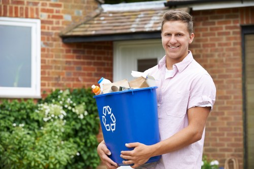 A young man with short, light brown hair is standing outside in front of a brick residential building with a tiled roof and a white-framed window. He is smiling and wearing a light pink, short-sleeved shirt while holding a large blue recycling bin filled with assorted rubbish, including paper, cardboard, plastic bottles, and mixed waste. The background features green shrubbery and potted plants along the paved driveway, indicating a suburban or urban residential area. The scene suggests a rubbish collection or clearance activity, performed by companies such as Office Clearance Shoreditch, serving areas like Shoreditch in London, with the bin positioned centrally in the image, and the man standing slightly to the right, engaging directly with the camera and fostering a professional, yet approachable atmosphere.