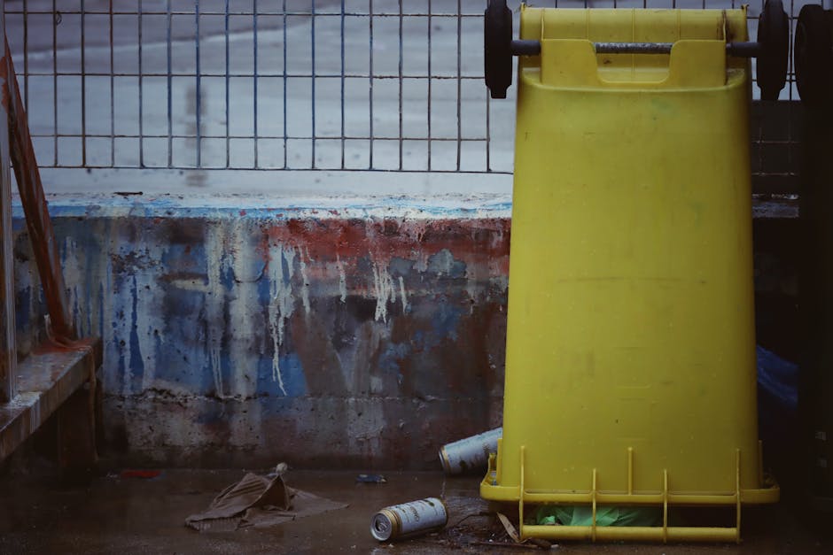 Two green wheeled rubbish bins with domed lids are positioned side by side on a paved sidewalk, close to the edge of the street. The bins are made of plastic with a smooth, slightly glossy finish and 