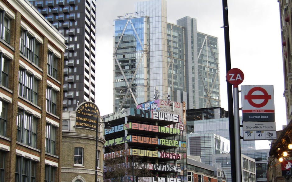 A black, cylindrical metal trash bin with a slightly textured surface and various stickers and graffiti tags affixed to it, situated on a concrete platform. The bin is partially covered by a black pla
