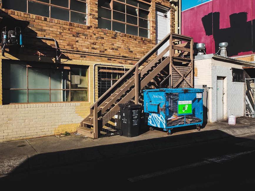 A row of large, rectangular wheelie bins lined up along a paved pathway adjacent to a brick wall. The bins are made of durable plastic with textured lids, and are colored green, blue, red, and dark gr