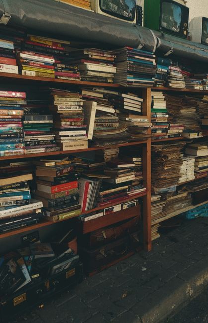 A large open-fronted wooden shelving unit located outdoors on a paved surface, filled with a diverse collection of books, magazines, and newspapers. The books vary in size, thickness, and binding mate