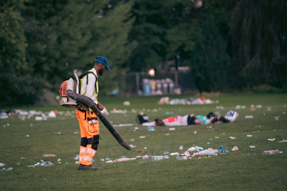 A man dressed in high-visibility orange and black trousers with a white high-visibility jacket and a blue cap is operating a leaf blower or vacuum device on a grassy field, surrounded by scattered lit