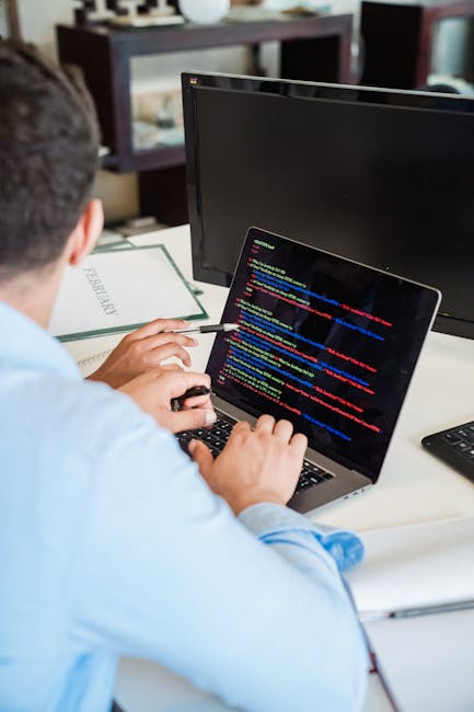 A black laptop with a glowing red and cyan keyboard displaying lines of blurred code on a dark screen, positioned on a flat surface. To the right of the laptop, a white Puma baseball cap with the blac